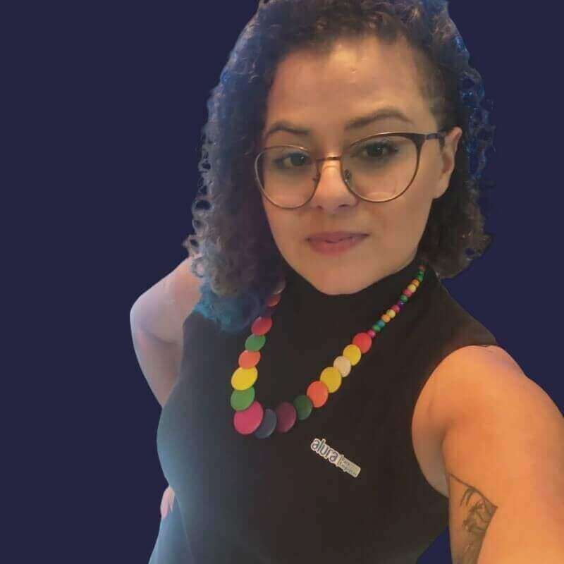 Mulher branca de cabelos cacheados e escuros, usando camisa da Alura Para Empresas, óculos pretos e colar colorido.